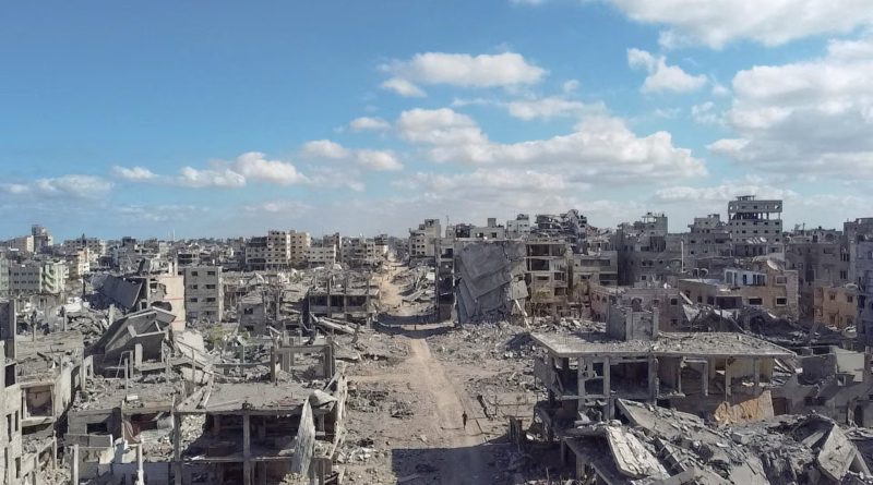 Wide view of Gaza City on October 18 showing heavily damaged and collapsed buildings lining a debris-covered road beneath a blue sky.